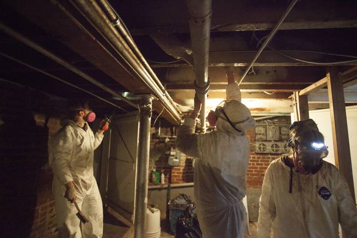 In a dimly lit basement. three volunteers in white coveralls and respiratory masks work, with large brushes, to remove mold from the ceiling.